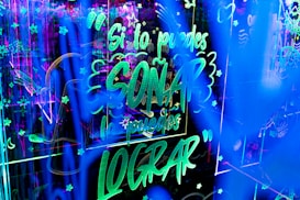 Bright neon lights create an energetic and lively atmosphere with glowing text and decorative symbols filling the space. The text, in Spanish, conveys an inspirational message amidst a backdrop of stars and abstract patterns.