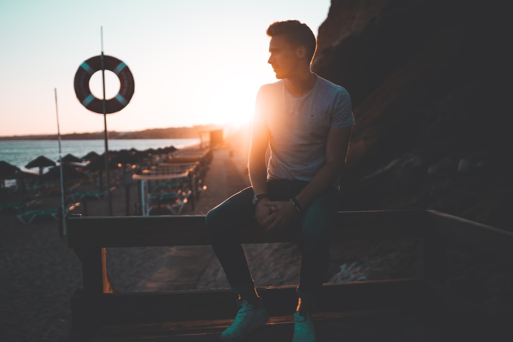 man in white crew neck t-shirt and black pants sitting on brown wooden bench during