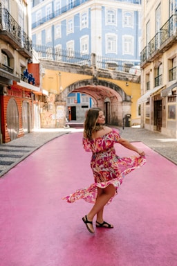 A woman in a floral dress is gracefully dancing on a pink street in an urban setting. Surrounding her are colorful buildings, including a striking blue one with white-trimmed windows. The atmosphere is relaxed and vibrant.