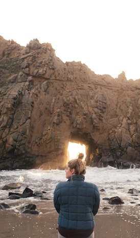 man in blue and black jacket sitting on rock formation in front of sea during daytime