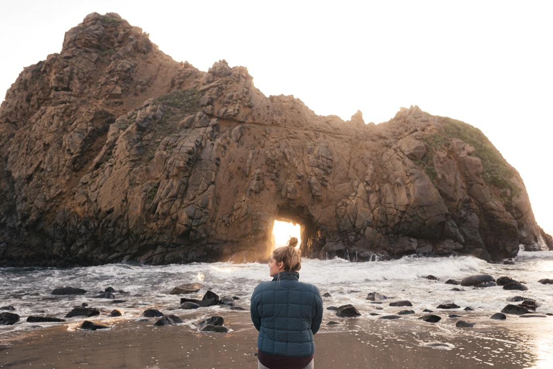 man in blue and black jacket sitting on rock formation in front of sea during daytime, Key hole rock<p></figure>
