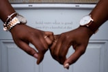 Two hands with watches are gently holding each other's pinkies in front of a white surface. Behind them, a motivational quote reads, 'hold onto what is good even if it's a handful of dirt.'