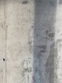 A textured concrete wall with visible signs of wear and a vertical shadow cast across part of its surface. The wall displays a rough texture with small holes and marks from the construction process.