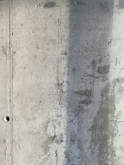 A textured concrete wall with visible signs of wear and a vertical shadow cast across part of its surface. The wall displays a rough texture with small holes and marks from the construction process.
