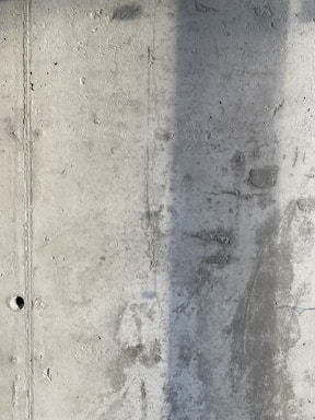 A textured concrete wall with visible signs of wear and a vertical shadow cast across part of its surface. The wall displays a rough texture with small holes and marks from the construction process.