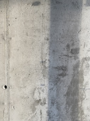 A textured concrete wall with visible signs of wear and a vertical shadow cast across part of its surface. The wall displays a rough texture with small holes and marks from the construction process.