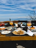 A wooden dining table is set with a variety of breakfast items and drinks against a backdrop of a scenic view. The table includes glasses of orange and pink drinks, plates with slices of bread, pastries, and a bowl of salad. A French press coffee maker is visible in the center of the table. In the background, there is a clear blue sky with soft clouds and distant mountains.