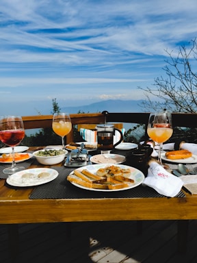 A wooden dining table is set with a variety of breakfast items and drinks against a backdrop of a scenic view. The table includes glasses of orange and pink drinks, plates with slices of bread, pastries, and a bowl of salad. A French press coffee maker is visible in the center of the table. In the background, there is a clear blue sky with soft clouds and distant mountains.