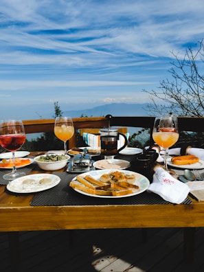 A wooden dining table is set with a variety of breakfast items and drinks against a backdrop of a scenic view. The table includes glasses of orange and pink drinks, plates with slices of bread, pastries, and a bowl of salad. A French press coffee maker is visible in the center of the table. In the background, there is a clear blue sky with soft clouds and distant mountains.