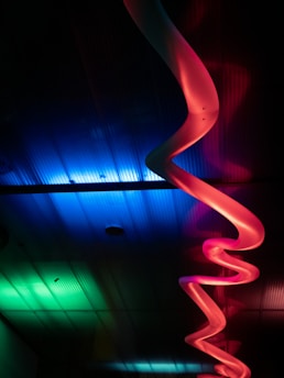 A colorful, abstract installation featuring a curving, ribbon-like structure illuminated by vibrant lighting. The structure is primarily red and pink, set against a backdrop of blue and green lights that create an ethereal atmosphere. The lighting casts soft shadows, adding depth and contrast to the scene.