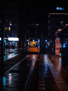 A tram approaches a station at night, illuminated by the glowing lights from nearby buildings and streetlights. The wet pavement reflects vivid colors, creating a vibrant atmosphere. Hotel signage is visible in the background, while a few people are scattered along the platform.