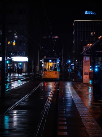 A tram approaches a station at night, illuminated by the glowing lights from nearby buildings and streetlights. The wet pavement reflects vivid colors, creating a vibrant atmosphere. Hotel signage is visible in the background, while a few people are scattered along the platform.