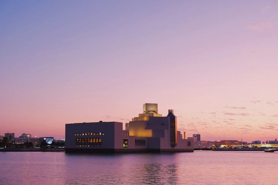 The image depicts a large white building with a dome-shaped roof, situated on an island in the middle of a body of water. The building is illuminated by warm orange lights and features multiple windows that reflect the surrounding environment. In the background, there are other buildings visible, suggesting that this island is part of a larger city or town. The sky above has a pinkish hue, indicating either sunrise or sunset, adding to the overall ambiance of the scene.