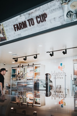 A coffee shop interior with a modern design, featuring a sign that reads 'Farm to Cup'. The counter is reflective, showing various coffee equipment and a barista. Behind the counter are shelves with colorful and neatly arranged products as well as a menu display.