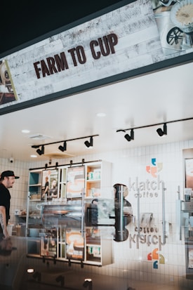 A coffee shop interior with a modern design, featuring a sign that reads 'Farm to Cup'. The counter is reflective, showing various coffee equipment and a barista. Behind the counter are shelves with colorful and neatly arranged products as well as a menu display.