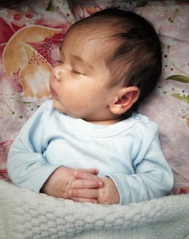An infant peacefully sleeps on a patterned floral blanket, wearing a light blue long-sleeved garment. The baby's hands are gently clasped together and tucked under a cozy white knit blanket.