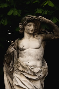 A classical stone statue depicting a partially draped figure with a muscular torso. The figure has a serene expression, curly hair, and is holding a cloth over its head. Lush green leaves frame the background, providing a contrast to the weathered texture of the statue.