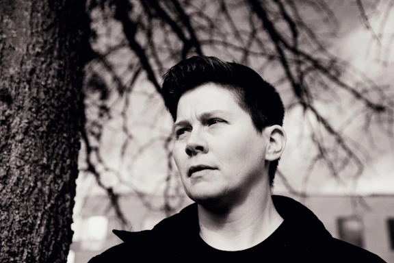 A black and white portrait of a person with short hair, gazing thoughtfully into the distance. The background features blurred tree branches and a building, imparting a sense of solitude and contemplation.