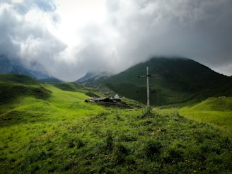 A serene, lush green landscape with rolling hills under a cloudy sky. A wooden cross stands prominently on a mound, and there is a small building nestled among the hills.