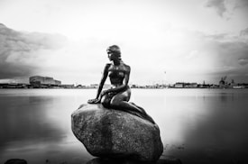 A statue of a mermaid sits gracefully on a rock by the waterfront. The background features an industrial cityscape with buildings and a calm, expansive body of water. The sky is cloudy, suggesting an overcast day.
