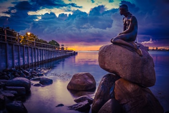 A statue of a mermaid sits gracefully on a large rock by a calm waterfront. The sky is filled with dramatic clouds, painted with shades of purple, blue, and orange from the setting sun. A pier lines the left side, adorned with lights that reflect on the water's surface.