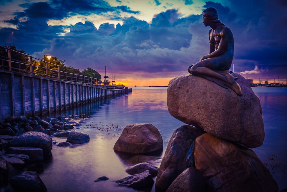 woman statue on rock near body of water during sunset