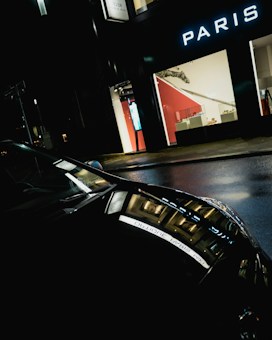 A dark street scene with reflections of bright storefront lights on a wet road and car. The storefront has the word 'PARIS' prominently displayed, and inside visible through the window are various display items. The overall atmosphere is moody and urban.