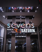 A modern storefront with a brightly illuminated sign. The sign reads 'sevens' with a playful, multicolored font, and 'home of SATURN' below in white. The interior showcases a store with elegantly designed products, and the surroundings have a sleek and luxurious appearance with visible brand signs like Emporio Armani.