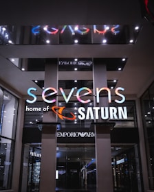 A modern storefront with a brightly illuminated sign. The sign reads 'sevens' with a playful, multicolored font, and 'home of SATURN' below in white. The interior showcases a store with elegantly designed products, and the surroundings have a sleek and luxurious appearance with visible brand signs like Emporio Armani.