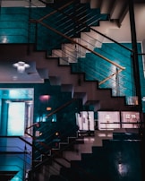 An indoor staircase with a modern design, featuring sleek metal railings and wooden handrails. The wall colors are a combination of teal and a neutral shade, creating a sophisticated atmosphere. The reflection of lights and a ceiling chandelier add an artistic and contemporary touch to the space.