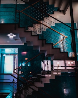 An indoor staircase with a modern design, featuring sleek metal railings and wooden handrails. The wall colors are a combination of teal and a neutral shade, creating a sophisticated atmosphere. The reflection of lights and a ceiling chandelier add an artistic and contemporary touch to the space.