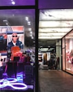 The image portrays a fashionable and modern store display with mannequins dressed in stylish clothing on the right side. The left side features a vibrant advertisement with a person in sunglasses and a leather jacket, surrounded by designer bags, highlighted by neon lighting.