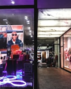 The image portrays a fashionable and modern store display with mannequins dressed in stylish clothing on the right side. The left side features a vibrant advertisement with a person in sunglasses and a leather jacket, surrounded by designer bags, highlighted by neon lighting.