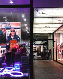The image portrays a fashionable and modern store display with mannequins dressed in stylish clothing on the right side. The left side features a vibrant advertisement with a person in sunglasses and a leather jacket, surrounded by designer bags, highlighted by neon lighting.