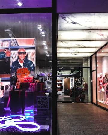 The image portrays a fashionable and modern store display with mannequins dressed in stylish clothing on the right side. The left side features a vibrant advertisement with a person in sunglasses and a leather jacket, surrounded by designer bags, highlighted by neon lighting.