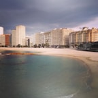 A coastal city scene with a line of modern apartment buildings facing a sandy beach. The sky appears overcast with dark clouds, and the water is calm and clear with varying shades of blue and green. There's a small patch of vegetation near the shoreline.