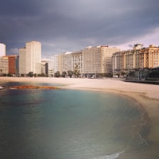 A coastal city scene with a line of modern apartment buildings facing a sandy beach. The sky appears overcast with dark clouds, and the water is calm and clear with varying shades of blue and green. There's a small patch of vegetation near the shoreline.