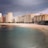 A coastal city scene with a line of modern apartment buildings facing a sandy beach. The sky appears overcast with dark clouds, and the water is calm and clear with varying shades of blue and green. There's a small patch of vegetation near the shoreline.