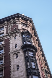 A detailed architectural facade of a historical building featuring ornate stone carvings, intricate cornices, and large windows with decorative frames. The structure primarily consists of red and beige bricks, accented by dark metal-windowed projections.