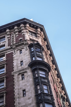 A detailed architectural facade of a historical building featuring ornate stone carvings, intricate cornices, and large windows with decorative frames. The structure primarily consists of red and beige bricks, accented by dark metal-windowed projections.