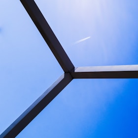 A contrail from an airplane is visible against a clear blue sky, framed by a geometric structure in the foreground.