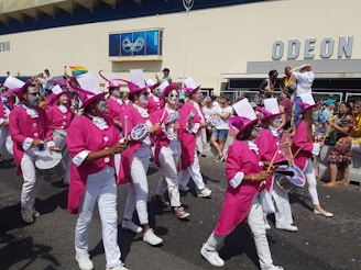 A group of people dressed in vibrant pink costumes and hats, with white face paint, are marching in a parade. They are playing drums and other percussion instruments. Spectators can be seen in the background, some holding flags or banners, and appear to be enjoying the event.