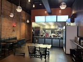 A cozy restaurant with a rustic brick wall on the left and a modern kitchen area visible at the back. The dining space features dark wooden tables and chairs, with booths lining the exposed brick wall. Pendant lights hang from the dark wooden ceiling, adding a warm ambiance. The kitchen area is brightly lit, showing two staff members preparing food behind a counter with a chalkboard menu.