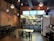 A cozy restaurant with a rustic brick wall on the left and a modern kitchen area visible at the back. The dining space features dark wooden tables and chairs, with booths lining the exposed brick wall. Pendant lights hang from the dark wooden ceiling, adding a warm ambiance. The kitchen area is brightly lit, showing two staff members preparing food behind a counter with a chalkboard menu.