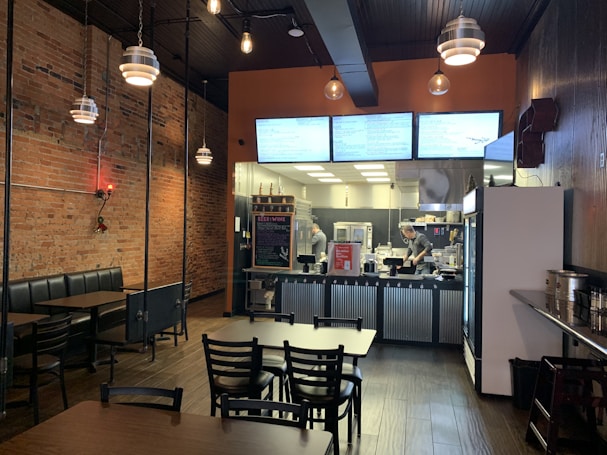 A cozy restaurant with a rustic brick wall on the left and a modern kitchen area visible at the back. The dining space features dark wooden tables and chairs, with booths lining the exposed brick wall. Pendant lights hang from the dark wooden ceiling, adding a warm ambiance. The kitchen area is brightly lit, showing two staff members preparing food behind a counter with a chalkboard menu.