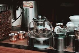 A glass teapot sits on a metal warmer surrounded by various containers and cups. On the left is a jar filled with coffee beans. Metallic cups with intricate designs are placed nearby, alongside a modern coffee maker. A serene and organized coffee-making setup is evident.