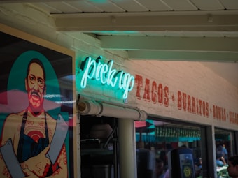 A vibrant neon sign reading 'pick up' glows in blue and green hues against a brick wall. Next to the sign is a colorful, stylized mural of a man holding two large knives, wearing an apron and showcasing tattoos. The brick wall has additional text promoting tacos, burritos, bowls, and salads.
