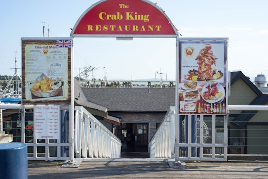 An entrance to the Crab King Restaurant featuring a large red sign above two menu boards. The left board displays a take-out menu, complete with images of fish and chips, while the right board shows various seafood platter options. The restaurant appears to be located near a waterfront, as indicated by visible fishing boats in the background.