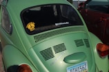 A green Volkswagen Beetle is parked next to a red car. The car displays a sticker on the rear window reading 'Powered by Love' with a peace sign and another message saying '0-60 Eventually!'. An Arizona license plate is visible with the number RTH1562.