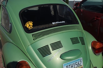 A green Volkswagen Beetle is parked next to a red car. The car displays a sticker on the rear window reading 'Powered by Love' with a peace sign and another message saying '0-60 Eventually!'. An Arizona license plate is visible with the number RTH1562.